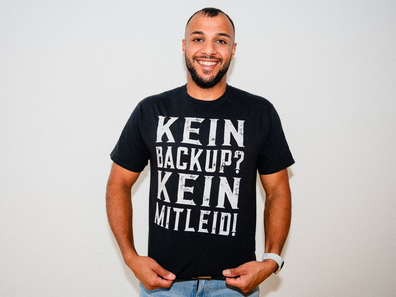 Ein lächelnder Mann mit kurzem Bart und kurz geschnittenem Haar steht vor einer weißen Wand. Er trägt ein schwarzes T-Shirt mit weißem, großgedrucktem Text: „KEIN BACKUP? KEIN MITLEID!“. Der Mann hält mit beiden Händen den unteren Rand seines T-Shirts leicht nach vorne gezogen. Er trägt eine helle Jeans und eine weiße Armbanduhr am linken Handgelenk. Die Stimmung ist freundlich und locker.